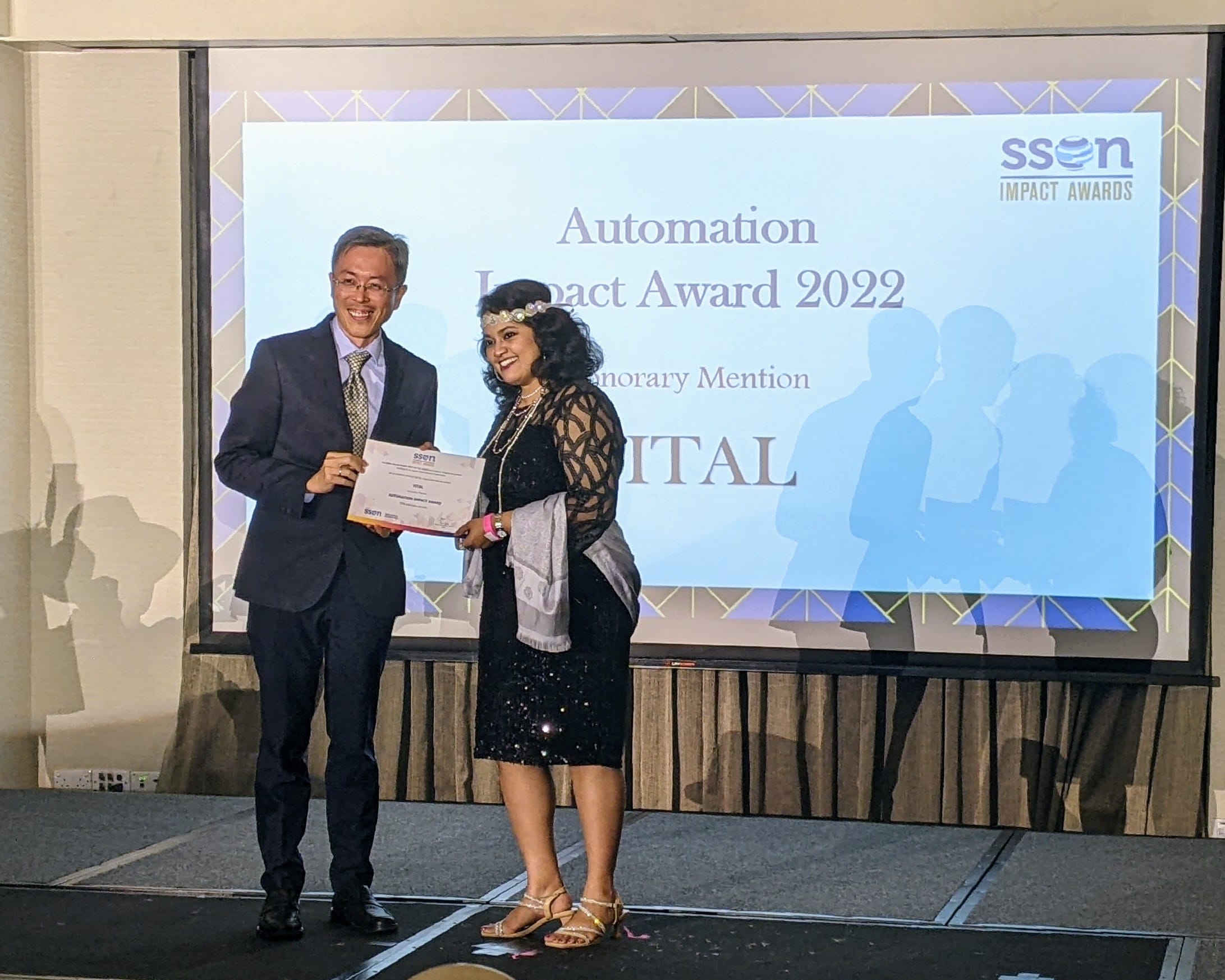 Two people stand on stage holding a certificate in front of a screen showing SSON Automation Impact Award 2022.